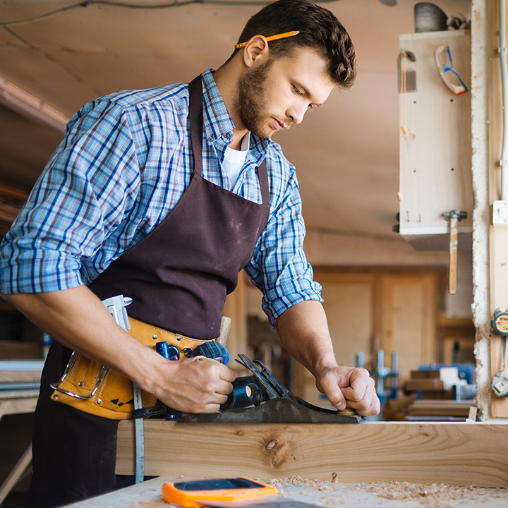 Profile view of confident young carpenter in apron planing board with jointer plane in spacious workshop